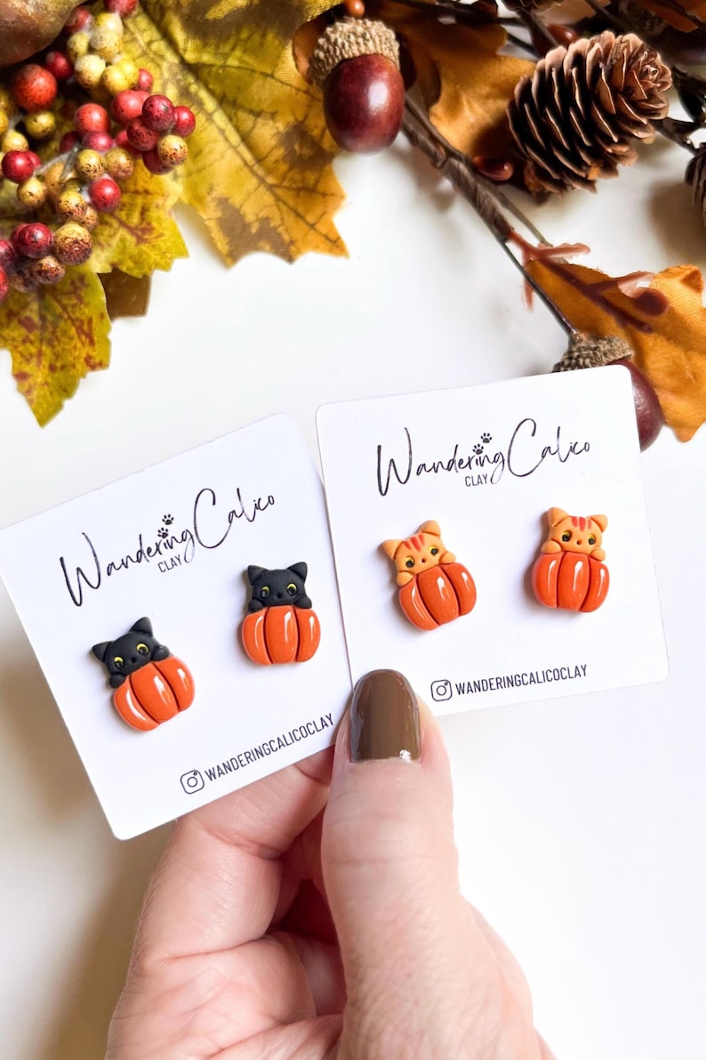 May include: Two pairs of handmade clay earrings featuring a black cat and a tabby cat inside orange pumpkins. The earrings are displayed on white cards with the text "Wandering Calico Clay" and social media handles. Autumn-themed jewelry.