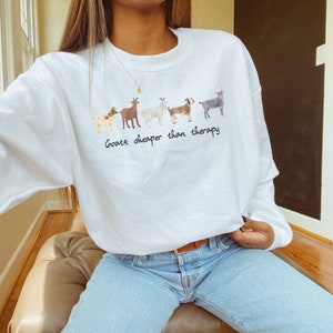 Op de afbeelding: Witte trui met een rij geiten bedrukt op de voorkant. De tekst "Goats: cheaper than therapy" is onder de geiten gedrukt.