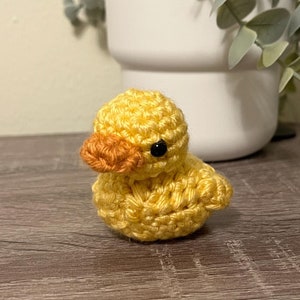 May include: A small crocheted yellow duck with an orange beak and black eyes. The duck is sitting on a wooden surface.
