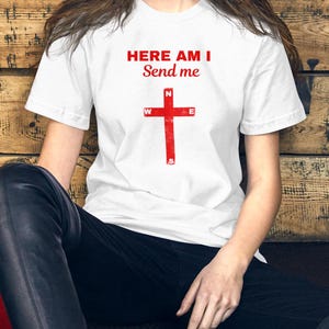 以下が含まれることがあります： 赤い文字で「HERE AM I Send me」と赤い十字架がプリントされた白いTシャツ。半袖のシャツは綿のようです。モデルは黒いパンツを着用しています。