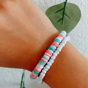 Pode incluir: Duas pulseiras em um pulso. Uma pulseira é branca com pequenas contas redondas. A outra pulseira é feita de contas redondas, planas e coloridas em tons de rosa, azul e branco.