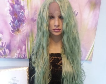 May include: A long, wavy, green wig with blonde highlights. The wig is styled with loose waves and is worn on a mannequin head.