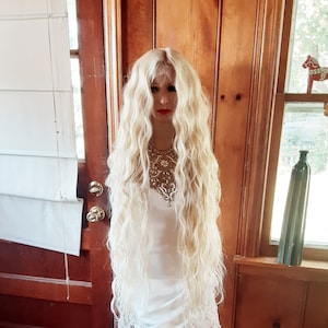 May include: A long, wavy, platinum blonde wig on a white mannequin head. The wig is styled in loose waves and falls to the mannequin's waist. The mannequin is wearing a white dress and is standing on a white stool.