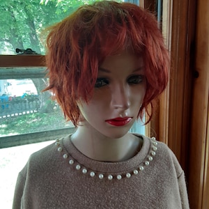 May include: A mannequin head with a short, layered, reddish-orange wig. The mannequin wears a tan sweater with a pearl necklace around the neckline. The mannequin has red lipstick and is positioned in front of a window.