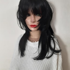 May include: A black wig with a layered, shaggy style and bangs. The wig is styled on a mannequin head with red lipstick and a white sweater. The wig has long, dark strands that frame the face.