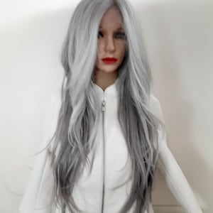 Puede incluir: Una peluca larga de color gris plateado con un estilo ondulado. La peluca está peinada con raya lateral y cae hasta los hombros. La peluca se lleva en una cabeza de maniquí con una chaqueta blanca.