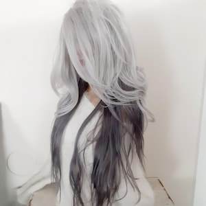 May include: A long, straight, two-toned wig with silver and dark gray hair. The wig is styled with loose waves and is worn by a person with a white sweater.