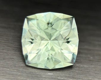 May include: A light green gemstone with a square cushion cut. The gem has many facets that reflect light, creating a sparkling effect. The stone is set against a dark background.