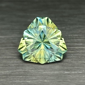 May include: A faceted gemstone with a triangular shape. The gem features a blend of yellow and blue-green hues, with a starburst pattern emanating from the center. The facets create a play of light and shadow.