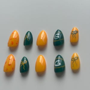 May include: A set of ten artificial nails in various designs. Some nails are yellow with orange spots, others are green with a yellow bow, and some are yellow with a green bow. Two nails have a green background with a white "G" logo.