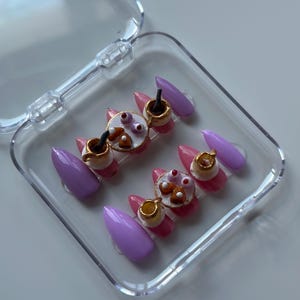 May include: A set of press-on nails in a clear plastic case. The nails feature a light pink base with gold accents, miniature teacups, and purple almond-shaped nails. The nail art includes tiny pastries and coffee cups.