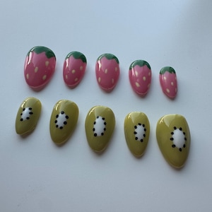 May include: A set of ten artificial nails, designed with fruit-themed patterns. Five nails feature a pink strawberry design with green tops, and the other five showcase a kiwi design with green and black details. The nails are oval-shaped.