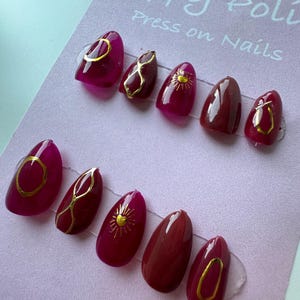 May include: A set of press-on nails in various shades of red and burgundy, with gold accent designs. The designs include geometric shapes and sunburst patterns. The nails are arranged on a light purple card with the text "Poppy Poli Press on Nails".
