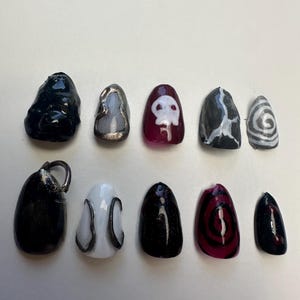 May include: A collection of ten artificial nails with various designs. The nails feature colors like black, white, silver, and burgundy. Some nails have skull, swirl, and abstract patterns. The nails are oval-shaped and appear to be made of acrylic or gel.