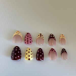May include: A set of ten artificial nails in various colors and designs. The nails feature a glossy finish and are decorated with polka dots. Colors include pink, yellow, and burgundy. Some nails have a nude base with a colored tip.