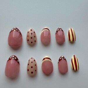 May include: A set of ten artificial nails in various designs. The nails feature a pink base with designs including red polka dots, red and white tips, and yellow and red stripes. The nails are oval-shaped.