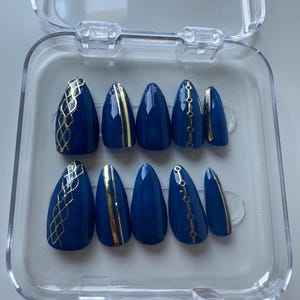 May include: A set of ten navy blue press-on nails with gold accents. The nails are almond-shaped and feature various gold designs, including geometric patterns and vertical stripes. The nails are displayed in a clear plastic case.