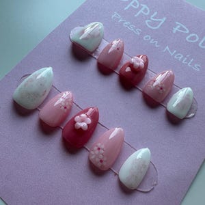 May include: A set of Poppy Pol press-on nails in various shades of pink and white. Some nails feature floral designs, while others have glitter accents. The nails are arranged on a lavender card with the brand name and product type printed on it.