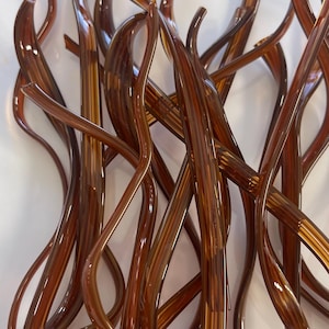 May include: A collection of long, slender, translucent brown glass rods with a smooth, glossy finish. The rods are curved and twisted, overlapping each other in a dense, tangled arrangement, creating a complex, multi-layered visual pattern.