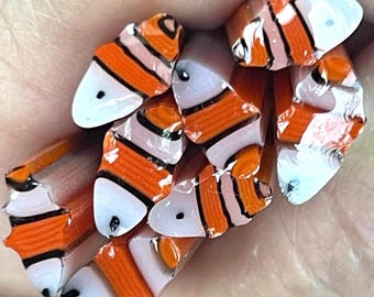 May include: A collection of small, cylindrical glass cane pieces featuring a repeating clownfish pattern. Each piece displays vibrant orange, white, and black stripes with a distinct eye detail, showcasing the intricate cross-section design.