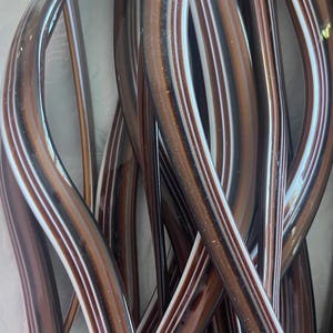 May include: A collection of long, slender, curved glass rods featuring a smooth, glossy finish. The rods display a layered color pattern of deep brown, translucent amber, and thin white stripes running vertically along their entire length.