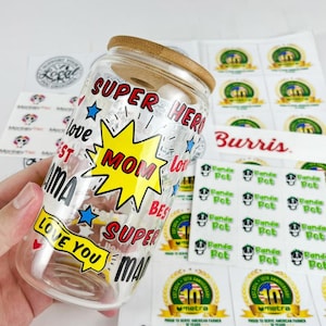 May include: A clear glass jar with a bamboo lid, decorated with the words "SUPER MOM" in a yellow burst, surrounded by "SUPER HERO" and "LOVE YOU." Additional stickers are visible in the background.