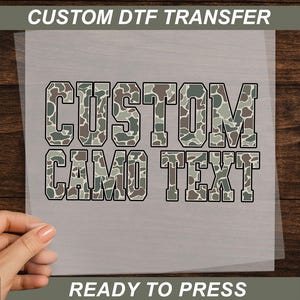 May include: A custom DTF transfer sheet with the words "CUSTOM CAMO TEXT" in a camouflage pattern. The text is outlined in black. The words "READY TO PRESS" are at the bottom.