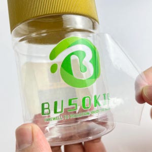 May include: A clear plastic jar with a gold lid and a green and white logo. The logo features a stylized letter "B". The text "BUSOKI" and "FAREWELL TO POLLUTION. ENJOY THE" are printed on the jar.
