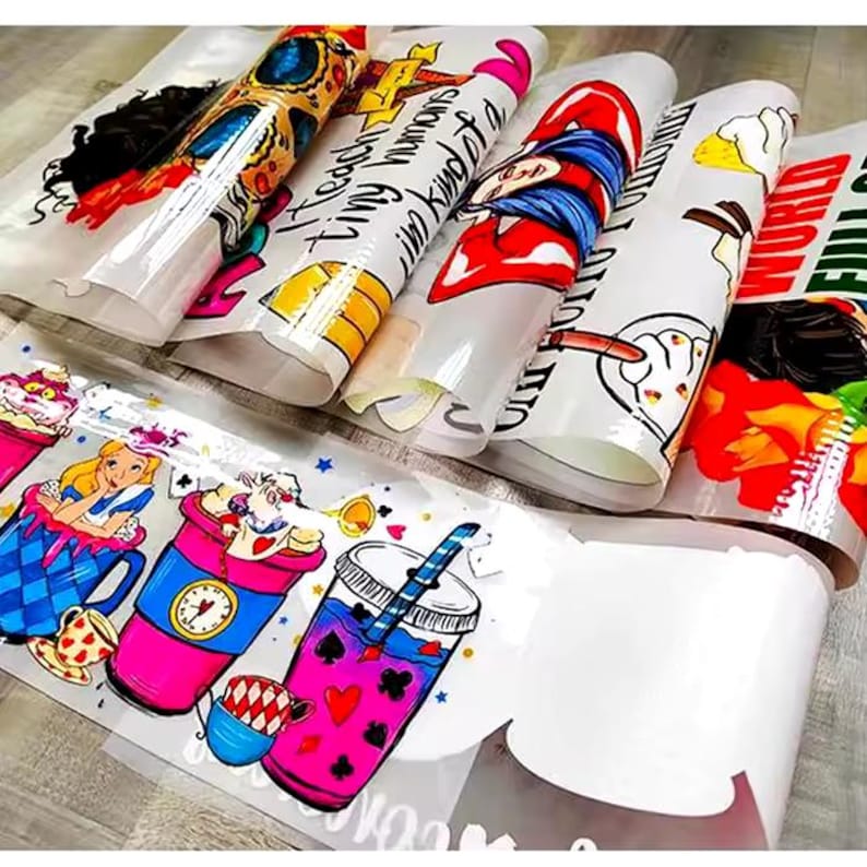 May include: Several rolls of printed vinyl sheets featuring colorful illustrations and text. Designs include Alice in Wonderland characters, cartoon figures, and phrases like "Teach tiny humans I'm kind of a big deal." The rolls are partially unrolled, revealing the printed designs.