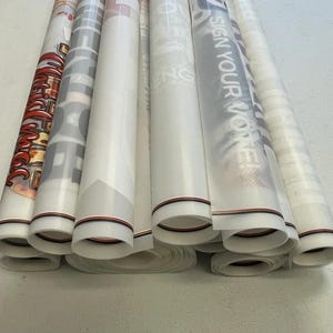 May include: A stack of rolled up white vinyl banners with black and red trim. The banners have various designs and text, including "Sign Your Vote" and "The Future is Now".