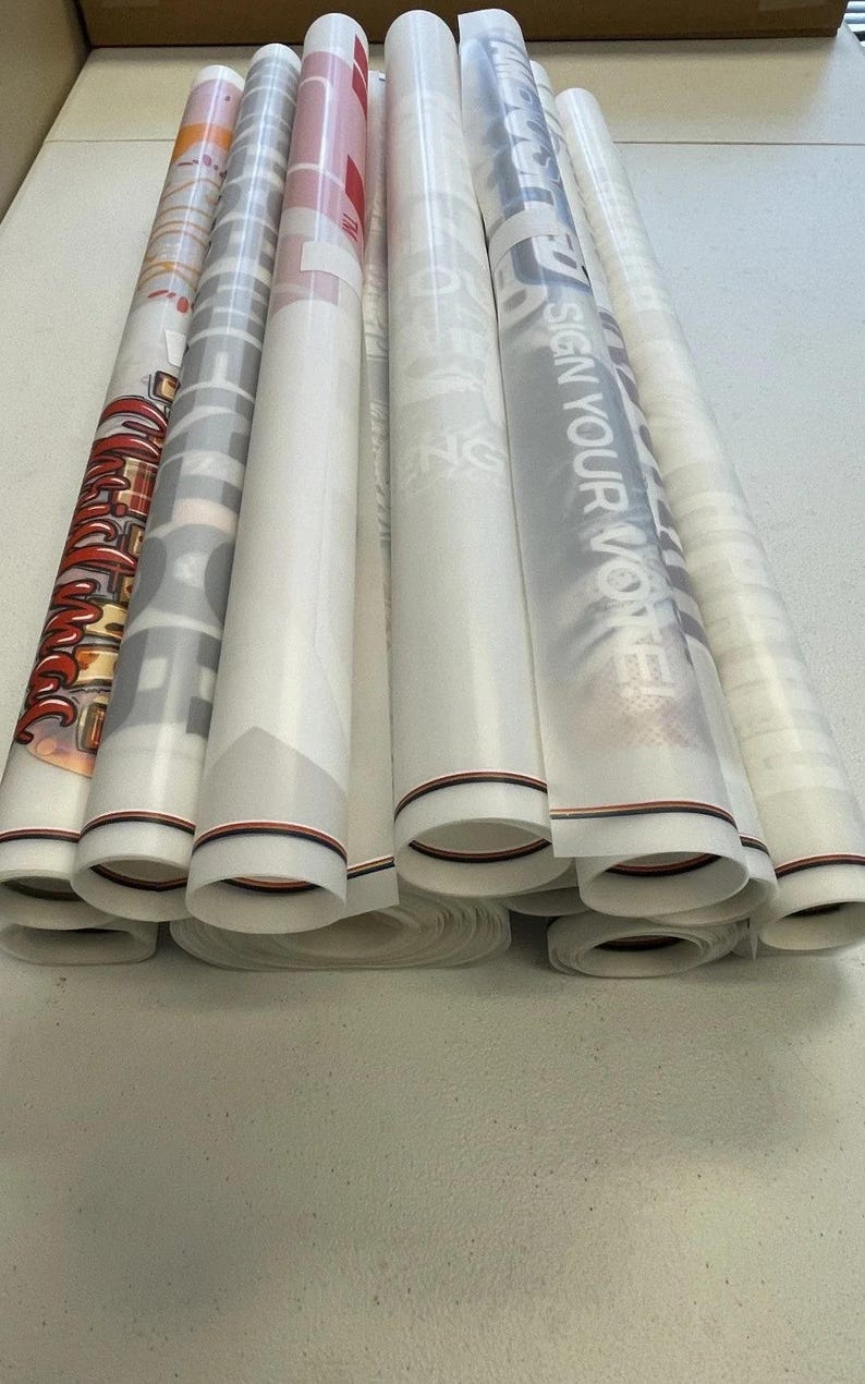 May include: A stack of rolled up white vinyl banners with black and red trim. The banners have various designs and text, including "Sign Your Vote" and "The Future is Now".