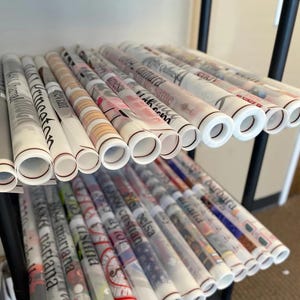 May include: A shelf filled with rolls of white wrapping paper with various names printed on them in black lettering. The rolls are stacked on top of each other and are arranged in a neat and organized manner.