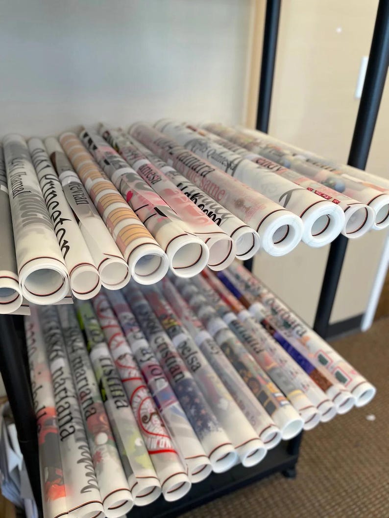 May include: A shelf filled with rolls of white wrapping paper with various names printed on them in black lettering. The rolls are stacked on top of each other and are arranged in a neat and organized manner.