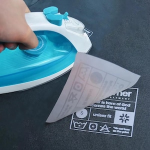 May include: A white and blue iron being used on a dark gray t-shirt. A white paper with care instructions and the text "mer" and "er is born of God comes the world" is being peeled back. The shirt has a small "S" and the words "unisex fit".