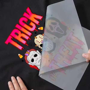 May include: Black fabric with a Halloween-themed design. The word "TRICK" is in pink and orange, with images of Halloween masks and candy corn. A clear sheet with the word "TREAT" is being peeled back.