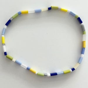 Op de afbeelding: Een kleurrijke armband gemaakt van kleine, rechthoekige kralen in tinten blauw, geel, groen en wit.