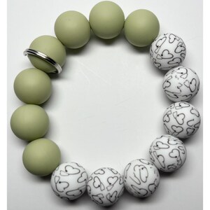 May include: A circular bracelet featuring alternating beads. The bracelet includes smooth, olive-green spherical beads and white beads adorned with black heart-shaped designs. A silver-colored metal ring is also present.