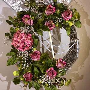 May include: A pink and green floral wreath with white pearl accents. The wreath is displayed on a gold metal stand.