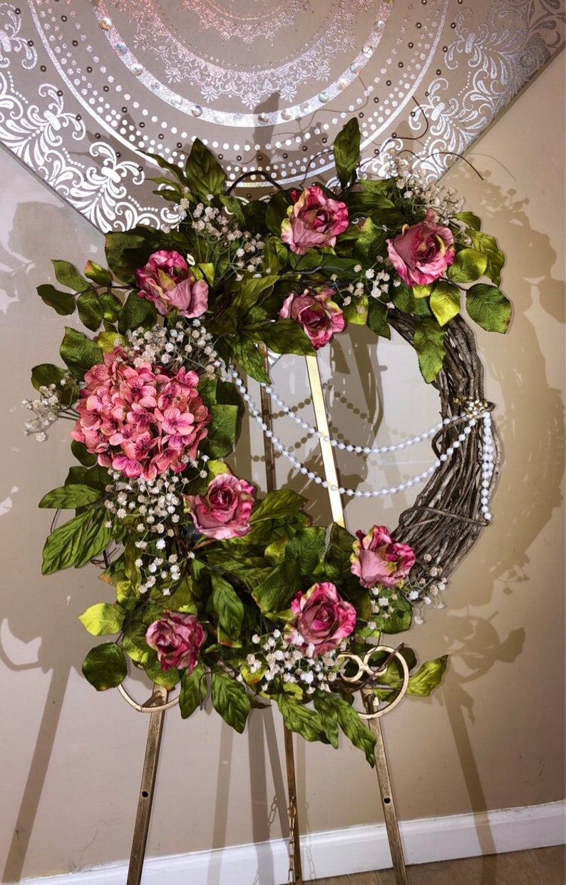 May include: A pink and green floral wreath with white pearl accents. The wreath is displayed on a gold metal stand.