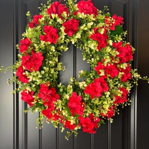 May include: A vibrant wreath featuring bright red flowers and lush green foliage. The wreath is circular and appears to be made of artificial materials. It is displayed on a black door, creating a striking contrast.