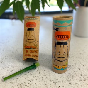 Puede incluir: Un tubo de cartón marrón con un gráfico colorido de una cabeza tiki y el texto "Kone-Tiki 5 Cones King Size". El tubo está al lado de un tubo de cartón marrón más pequeño con un gráfico similar y el texto "Kone-Tiki".