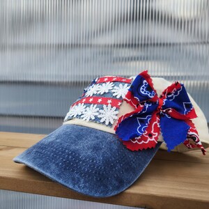 May include: A baseball cap featuring a distressed blue denim brim and a cream-colored crown. The front panel is decorated with a rectangular patch of blue denim, white floral lace, and red ribbon with white stars. A large red and blue bandana bow is attached.