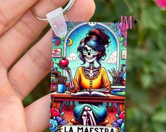 Könnte beinhalten: Ein rechteckiger Schlüsselanhänger mit einer farbenfrohen Illustration einer Skelettlehrerin mit Brille, die einen Apfel hält und an einem Schreibtisch sitzt. Der Text "LA MAESTRA" steht unten. Der Schlüsselanhänger hat einen silbernen Ring.