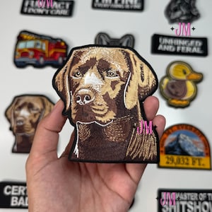 May include: A selection of embroidered patches with diverse designs. The main patch depicts a detailed brown Labrador Retriever head. Other patches feature text such as "I'M FINE" and "UNHINGED AND FERAL", and a mountain scene.