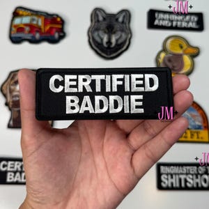 May include: A collection of embroidered patches with various designs and text. One patch reads "CERTIFIED BADDIE" in white on a black background. Other patches include a wolf, a duck, and the text "FUN FACT I DON'T CARE".