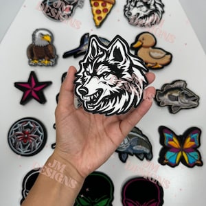 May include: A collection of iron-on patches featuring various designs, including a wolf, a skull, a spider, a butterfly, a fish, a duck, a pizza slice, a star, and an eagle. The patches are all colourful and detailed.
