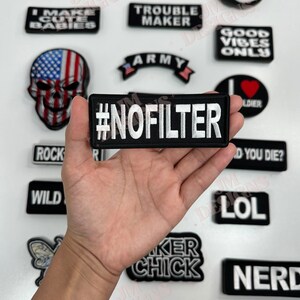 May include: A hand holding a black and white iron-on patch with the text "#NOFILTER" in white letters. The patch is surrounded by other iron-on patches with various designs and text, including "I Make Cute Babies", "No Thanks", "Trouble Maker", "Army", "Good Vibes Only", "Soldier", "Rock", "Wild", "LOL", "Biker Chick", "Nerd", and "Biker Babe".