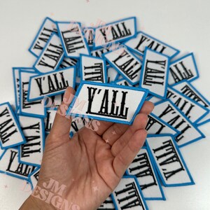 May include: A hand holding a blue and white iron-on patch with the word "Y'all" in black lettering. The patch is surrounded by a stack of similar patches.