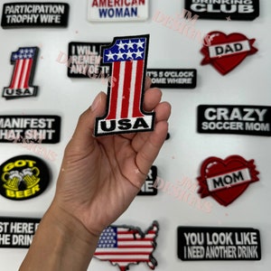 Könnte beinhalten: Eine Sammlung von Bügelbildern mit verschiedenen Designs, darunter ein rotes Herz mit dem Wort "DAD" darauf, ein rotes Herz mit dem Wort "MOM" darauf, ein schwarz-weißes Patch mit den Worten "CRAZY SOCCER MOM", ein schwarz-weißes Patch mit den Worten "MANIFEST THAT SHIT", ein schwarz-weißes Patch mit den Worten "MID DAY DRINKING CLUB", ein schwarz-weißes Patch mit den Worten "AMERICAN WOMAN", ein schwarz-weißes Patch mit den Worten "PARTICIPATION TROPHY WIFE", ein schwarz-weißes Patch mit den Worten "YOU LOOK LIKE I NEED ANOTHER DRINK", ein schwarz-weißes Patch mit den Worten "IT'S 5 O'CLOCK SOMEWHERE", ein schwarz-weißes Patch mit den Worten "I WILL REMEMBER ANY OF THIS", ein schwarz-weißes Patch mit den Worten "GOT BEER", ein schwarz-weißes Patch mit den Worten "ST HERE THE DRINK", ein schwarz-weißes Patch mit den Worten "USA" und einer rot-weiß-blauen amerikanischen Flagge, und ein schwarz-weißes Patch mit den Worten "USA" und einer rot-weiß-blauen amerikanischen Flagge in Form der Zahl eins.