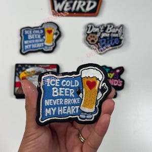 Op de afbeelding: Een set geborduurde strijkpatches met verschillende ontwerpen, waaronder een bierglas met een hart, een adelaar en een hondenpootafdruk. De patches zijn allemaal op een witte achtergrond en hebben verschillende kleuren, waaronder blauw, rood, geel en zwart. De tekst op de patches luidt "Ice Cold Beer Never Broke My Heart", "Weird", "Don't Bark If You Can't Bite", en "Friends".